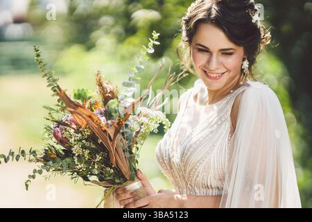 Schöne Braut mit Blumenstrauß Blumen steht auf Wald Hintergrund. Landhausstil. schöne Braut in zarten Kleid im Freien. Nahaufnahme, Porträt der jungen Stockfoto