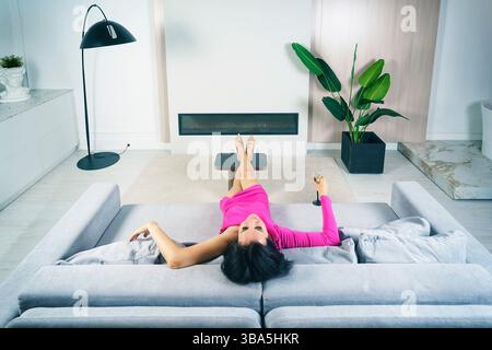 Eine hübsche Brünette in einem rosa Kleid liegt mit einem Glas Champagner auf dem Sofa Stockfoto