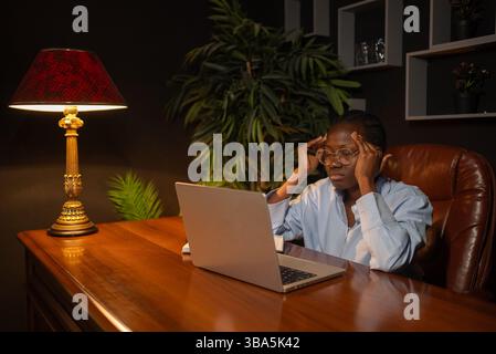 Professionelle afroamerikanische Arbeiterin, die unter intensivem Stress leidet und die Fingerspitzen gegen die Stirn drückt, während sie bis spät unter weichem Lampenlicht arbeitet, und der Laptop-Bildschirm wirft Schatten Stockfoto