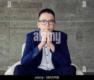 Ein Junge mit einer Brille mit intelligentem Look sitzt in einem Stuhl, die Hände sind in einem Schloss zusammengeklemmt Stockfoto