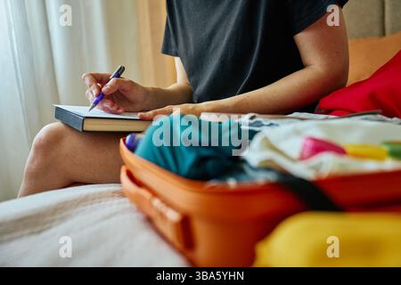 Die Person schreibt in einem Notizbuch mit einem orangen Koffer voller Kleidung. Reisekonzept mit handschriftlicher Checkliste Stockfoto