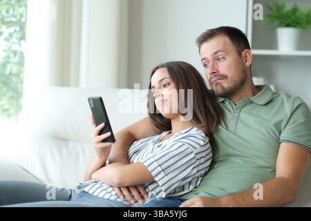 Enttäuschtes Paar, das zu Hause auf einer Couch auf langweiligen Online-Inhalten nachsieht Stockfoto