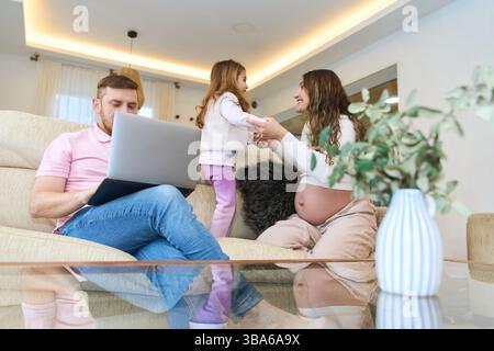 Glückliche schwangere Mutter, die mit ihrer Tochter auf dem Sofa spielt, während der Vater von zu Hause aus mit einem Laptop arbeitet und Familie, Arbeit und Pregn repräsentiert Stockfoto