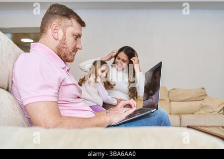 Vater benutzt Laptop mit schwangeren Frau und Tochter, die neben ihm auf der Couch sitzen und von zu Hause aus arbeiten Stockfoto