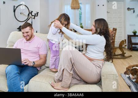 Glückliche schwangere Mutter spielt mit ihrer Tochter auf dem Sofa, während der Vater von zu Hause aus auf seinem Laptop arbeitet Stockfoto