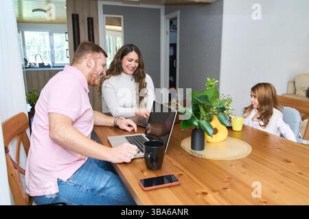 Glückliche lateinamerikanische Familie, die zu Hause Laptops benutzt, während die Tochter frühstückt, schwangere Mutter und Vater arbeiten von zu Hause aus Stockfoto
