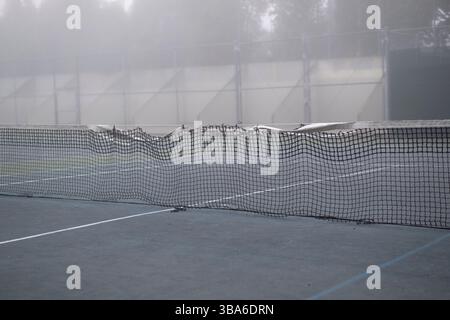 Beschädigtes Tennisnetz auf blauem Platz in dichtem, trübem Nebel Stockfoto