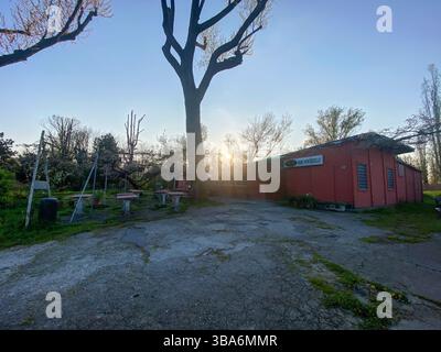 Castelvetro Piacentino, Italien - 5. April 2025 Sonnenaufgang hinter dem alten Holzgebäude unter einem Baum, der Hinweise auf die Natur an einem sonnigen Tag bietet Stockfoto