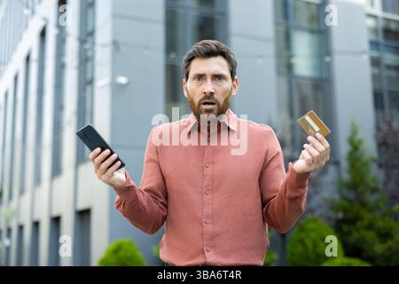 Ein Mann mit besorgtem Ausdruck hält eine Kreditkarte und ein Smartphone und sieht sich im Freien scheinbar mit finanziellen Problemen konfrontiert. Stockfoto