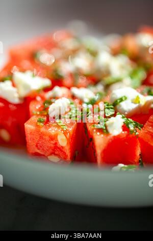 Reife Wassermelonenstücke, verstreut mit zerbröckelten Feta- und Minzblättern, eingebettet in eine hellgrüne Keramikschale vor tiefem schattigem Hintergrund Stockfoto