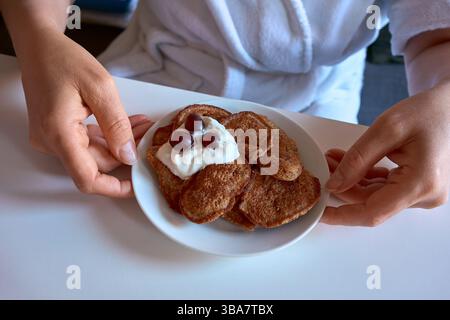 Frau in einem weißen Bademantel mit einem Teller gesunder hausgemachter Pfannkuchen mit Joghurt und Beeren. Gemütliches Frühstück, Lifestyle-Szene zu Hause. Stockfoto