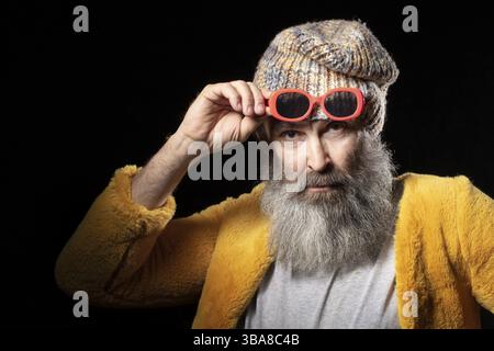 Ein älterer Mann mit grauem Bart, der einen Strickmütze und einen hellgelben Pelzmantel trägt. Modischer Mann auf schwarzem Hintergrund Stockfoto