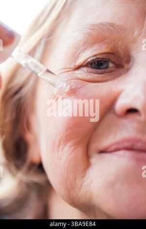 Nahaufnahme einer älteren Frau, die ein feuchtigkeitsspendendes Serum auf ihr Gesicht aufträgt. Reife Dame mit Pipette und Anti-Aging-Gel. Hautpflege im Erwachsenenalter Stockfoto