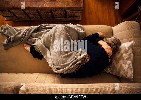 Eine ältere Frau liegt mit einer Decke bedeckt und leidet zu Hause an Kopfschmerzen. Eine reife Dame schläft auf der Couch und umarmt sich an den Schultern. Stockfoto