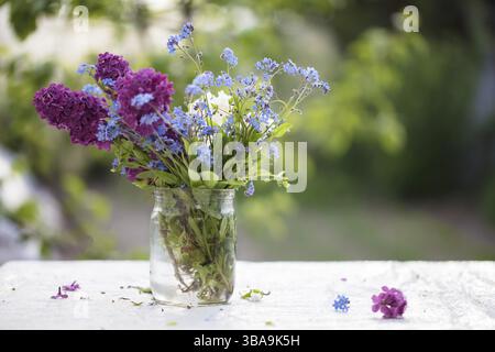 Frühling oder Sommer Hintergrund. Ein Blumenstrauß aus Flieder und Vergissmeinnots in einem Glas auf dem Tisch. Schöne Blumen auf grünem Hintergrund Stockfoto