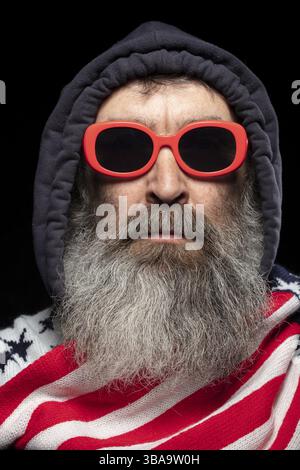 Ein älterer Mann mit grauem Bart, der eine Kapuze und einen Schal mit einem Muster der amerikanischen Flagge auf schwarzem Hintergrund trägt Stockfoto