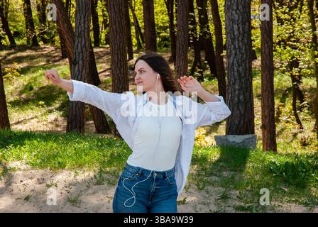 Junge kaukasische Frau, die Musik genießt, während sie in einem sonnendurchfluteten Wald tanzt, lässige Kleidung trägt und Freude und Freiheit in der Natur ausdrückt. Digital Nomad li Stockfoto