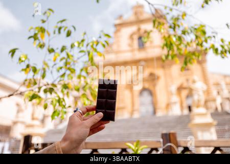 Nahaufnahme einer Hand mit traditioneller Modica-Schokoladentafel mit verschwommenem Blick auf die Kathedrale St. Peter in Modica, Sizilien, eine Hommage an den authentischen Geschmack und das Erbe Stockfoto