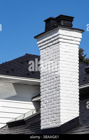 Schornstein auf dem Dach des weißen Hauses mit blauem Himmel im Hintergrund. Ein Wohndach mit einem weißen Ziegelkamin auf dem schwarzen Schindeldach. Architec Stockfoto