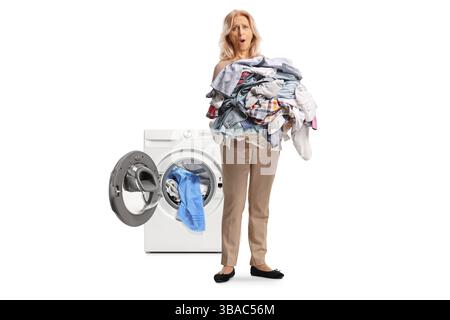 Wütende Frau, die einen großen Stapel Kleider vor einer Waschmaschine hält, isoliert auf weißem Hintergrund Stockfoto