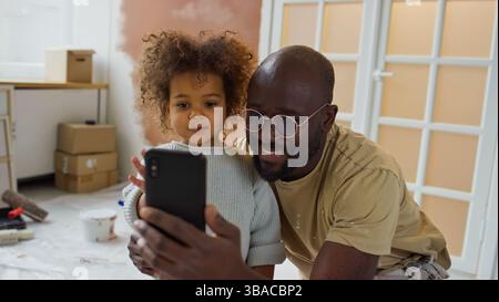 Vater, der mit seiner Tochter ein Selfie in einem renovierten Zimmer macht, zeigt ihre glücklichen Ausdrucksformen und ihre herzliche Verbindung mit einigen Renovierungsmitteln im Hintergrund Stockfoto