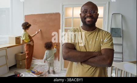 Porträt eines lächelnden afroamerikanischen Mannes mit überkreuzten Armen, der im renovierten Zimmer steht, mit zwei Kindern, die die Wand im Hintergrund malen Stockfoto