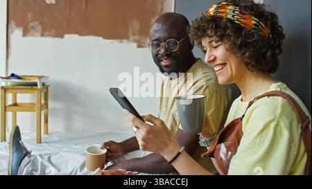 Afroamerikanischer Mann und kaukasische Frau sitzen auf dem Boden und halten Kaffeetassen und lächeln, während sie auf das Smartphone schauen. Farbausrüstung und Holzstuhl im Hintergrund Stockfoto