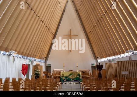 Das Innere einer modernen Kirche mit hoher Holzdecke, einem großen Kreuz an der Wand und einer Reihe von Holzstühlen. Der Altar ist mit einem Grün dekoriert Stockfoto
