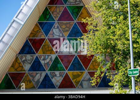 Ein farbenfrohes Buntglasfenster mit geometrischen Mustern und religiösen Figuren, vor einem Hintergrund von Grün und einem Straßenschild mit der Aufschrift „C“ Stockfoto