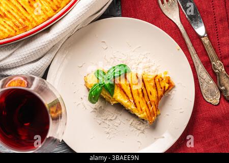 Auflauf mit Kartoffelpüree und Hackfleisch in einer Auflaufform und ein Stück Auflauf auf einem weißen Teller mit einer weinroten Serviette und Besteck auf grauem Hintergrund mit einem Glas Rotwein. Hochwertige Fotos Stockfoto