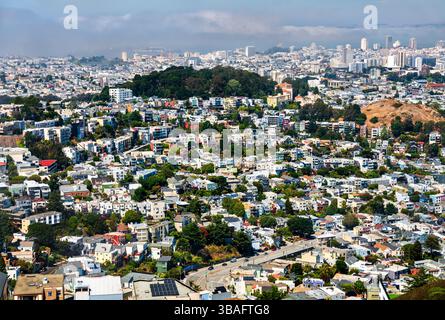 Dicht gepackte Wohnviertel von San Francisco, von oben betrachtet, mit farbenfrohen Häusern auf steilen Hügeln, Sutro Forest in der Mitte und einer nebelbedeckten Skyline im Hintergrund an einem klaren Sommertag. Stockfoto