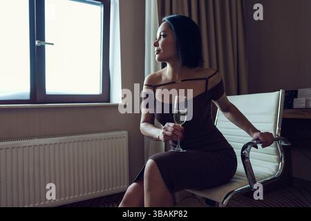 Eine dunkelhäutige Brünette sitzt nachdenklich auf einem Stuhl mit einem Glas Champagner in der Hand Stockfoto