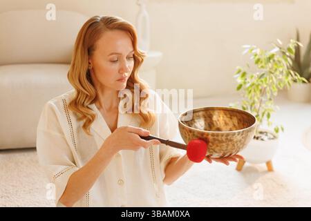 Ruhige Frau, die tibetische Klangschale für Klangtherapie und Meditation in einem friedlichen Innenraum mit sanftem Licht verwendet. Stockfoto
