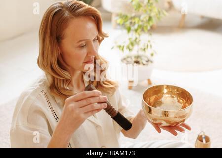 Friedliche Frau, die eine tibetische Klangschale benutzt, um Achtsamkeit in einem hellen, ruhigen Meditationsraum zu üben. Stockfoto