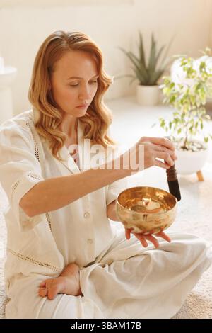 Frau, die während der Klangtherapie eine tibetische Klangschale benutzt und Achtsamkeit in einem beruhigenden Raum übt. Stockfoto
