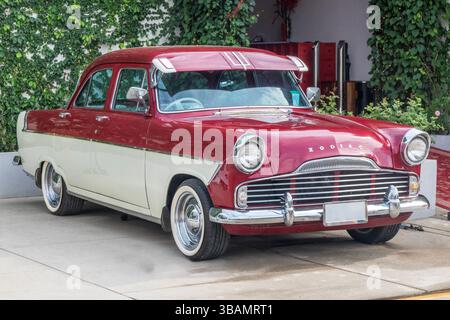 Ein klassischer zweifarbiger Ford Zodiac-Wagen in Rot und Creme parkt auf einer Betoneinfahrt neben einem mit grünen Kletterpflanzen bedeckten Gebäude. Der Vintage Stockfoto
