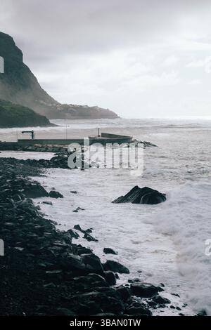 Dramatische Meereswellen stürzen gegen eine felsige Küste mit einer entfernten Stadt und einem Pier Stockfoto