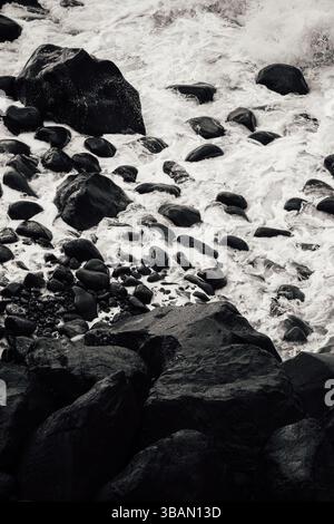 Dramatische Schwarzweiß-Nahaufnahme von Wellen, die über dunkle, nasse Küstenfelsen krachen Stockfoto