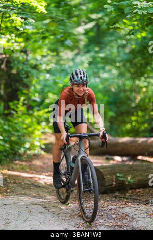 Lächelnde Radfahrerin in Sportkleidung und Helm fährt mit dem Schotterrad auf dem Waldweg. Konzept des Outdoor-Abenteuers, Radfahren durch die Natur, an Stockfoto