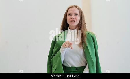 Eine junge Frau in einem grünen Blazer zeigt mit einem selbstbewussten Ausdruck, der Durchsetzungskraft und Entschlossenheit ausstrahlt. Der helle Hintergrund zeichnet den Fokus Stockfoto