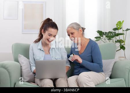 Die Enkelin hilft Großmutter, Seniorin und junge Frau beim Online-Einkaufen zu Hause mit Laptop und Kreditkarte auf dem Sofa. Konz Stockfoto