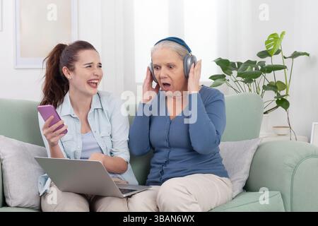 Ältere Frau reagiert überraschend, während sie Musik über kabellose Kopfhörer hört. Junge Frau lacht und hält Smartphone und Laptop. Spaß Stockfoto