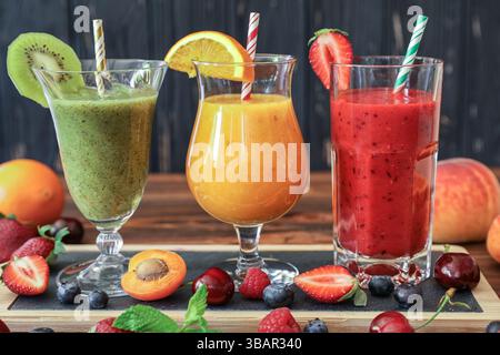 Drei große Gläser mit grünen, orangen und roten Smoothies sind auf einem Holztisch angeordnet, umgeben von frischem Obst wie Erdbeeren, Kiwis und Kirschen, perfekt für heiße Tage. Stockfoto