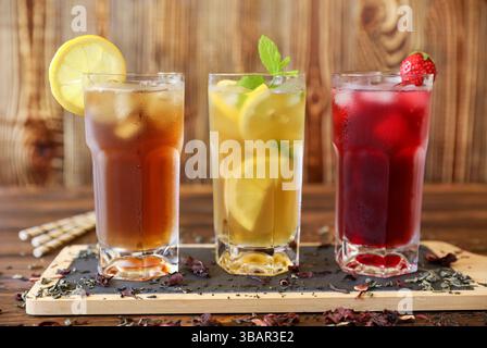 Auf einem rustikalen Holztisch stehen drei große Gläser mit Eistee, Limonade mit Zitronenscheiben und ein Beerengetränk mit Kirschen. Frische Minzgarnituren sorgen für einen lebendigen Touch. Stockfoto