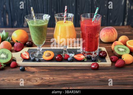 Drei große Gläser Smoothies in Grün-, Gelb- und Rottönen sitzen auf hölzernem Servierbrett. Die umliegenden Früchte umfassen Erdbeeren, Kiwi, Pfirsiche und Orangen, die eine erfrischende Atmosphäre schaffen. Stockfoto