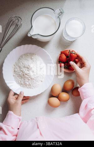 Die Zutaten zum Backen sind auf einer Küchentheke ordentlich angeordnet, einschließlich Mehl, Zucker, Milch, Eier, und frische Erdbeeren. Die Hände sind abgebildet, die einen halten Stockfoto