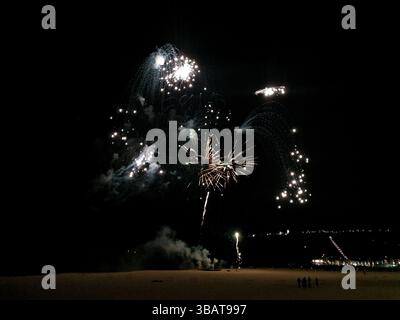 Spektakuläres Feuerwerk über einem dunklen Strand bei Nacht. Lebendige Lichtstöße erhellen den Himmel und schaffen eine dramatische und festliche Szene. Stockfoto