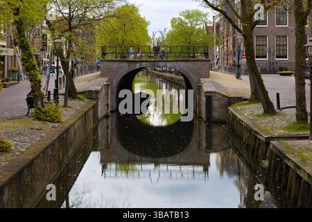Bogenbrücke über einen Kanal in Edam, Niederlande, mit Menschen, traditionellen Gebäuden und Bäumen, die sich im Wasser unter einem bewölkten Himmel spiegeln. Stockfoto