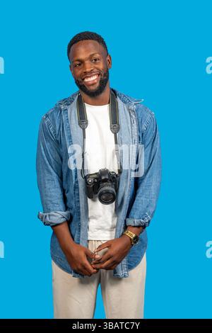 Junger Fotograf lächelt mit professioneller Kamera, die am Hals auf blauem Hintergrund hängt Stockfoto