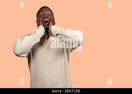 Überraschter Mann, der seine Wangen mit den Händen hielt und den Mund auföffnete, auf pfirsichfarbenem Hintergrund stehend Stockfoto